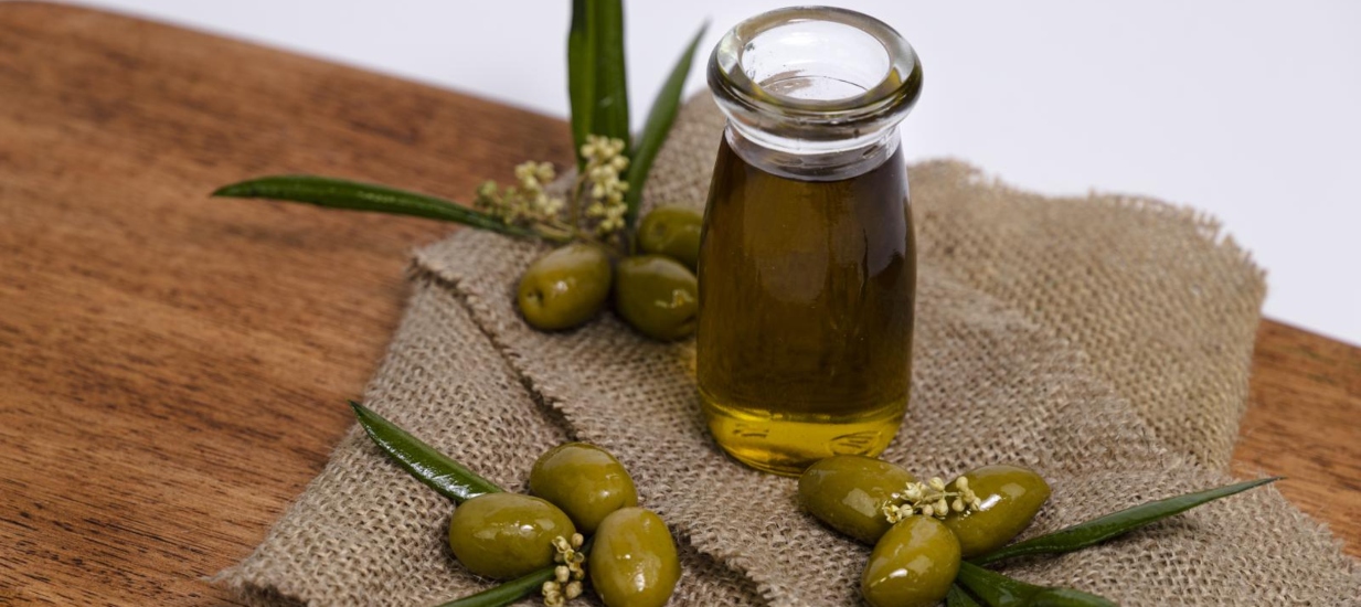 Botella de aceite de oliva virgen extra y aceitunas verdes frescas sobre tela de saco y tabla de madera, representando la calidad de los productos peruanos del sur