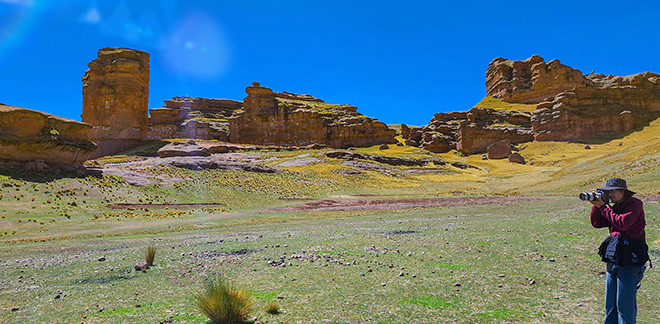Fotógrafo capturando la imponente belleza del Cañón de Tinajani en Puno, Perú. La escena muestra formaciones rocosas rojizas, un cielo azul intenso y una extensión de pastizal andino en este valle espiritual