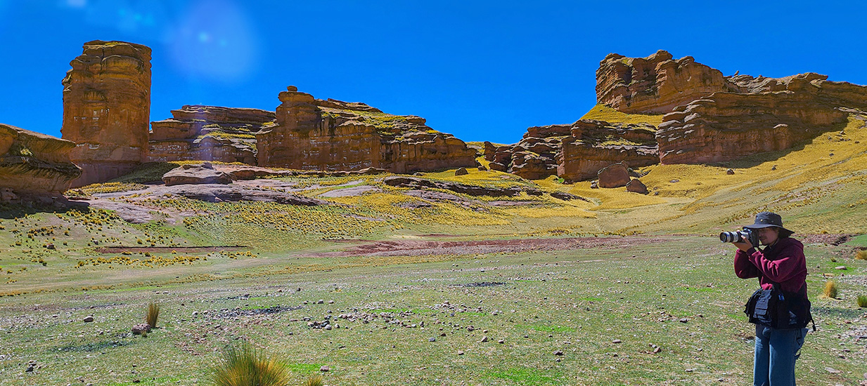 Fotógrafo capturando la imponente belleza del Cañón de Tinajani en Puno, Perú. La escena muestra formaciones rocosas rojizas, un cielo azul intenso y una extensión de pastizal andino en este valle espiritual