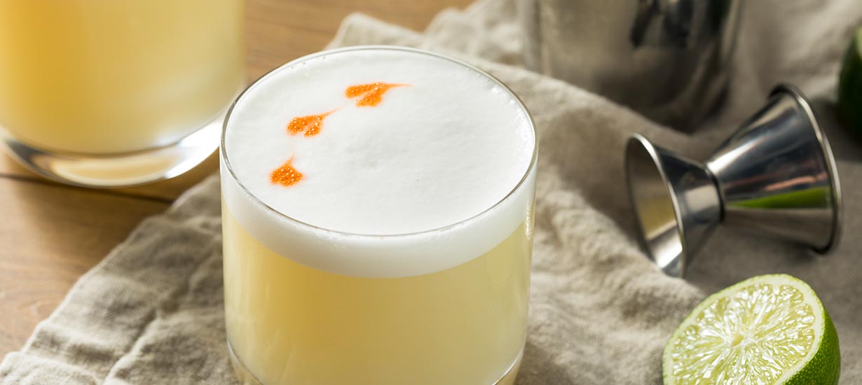 Pisco Sour en vasos de cóctel con espuma blanca, amargo y limas frescas.